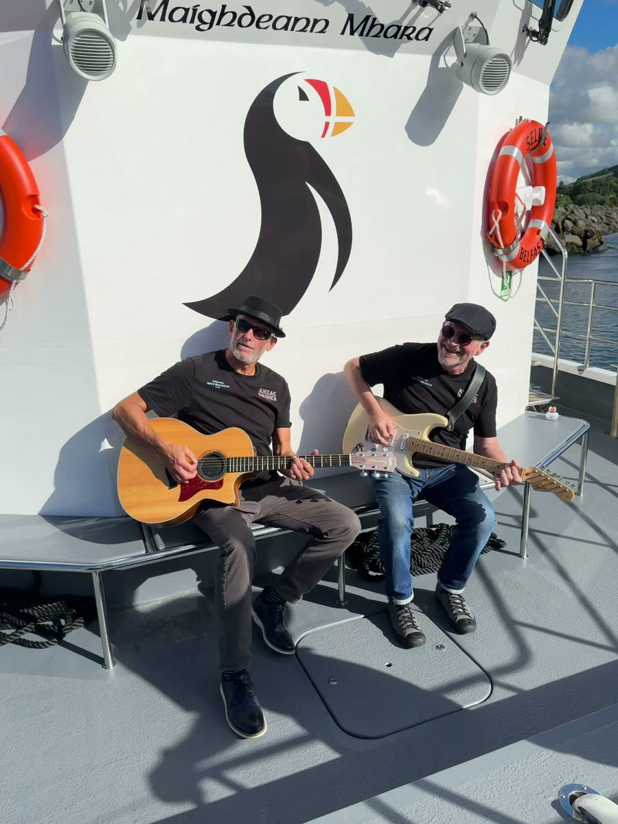 Two musicians sitting on the deck of Selkie playing guitars in the sun, the large Kintra puffin logo on the cabin behind them.