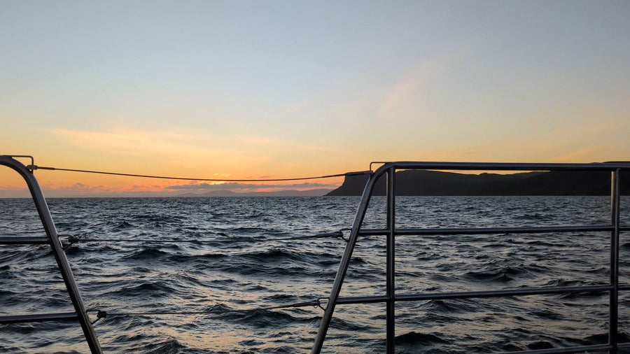 An orange sunrise sky over a dark sea with a headland on the horizon, seen past the rails of Selkie on the Darkness Into Light sunrise sailing from Ballycastle.
