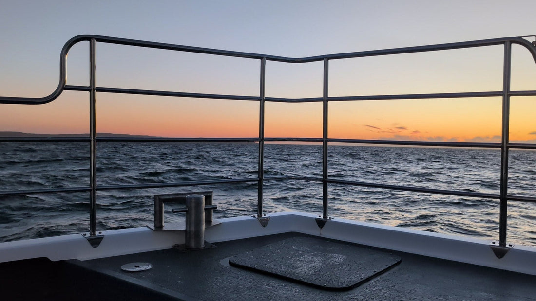 The bow rail of the boat Selkie in the foreground with an orange band of dawn light on the horizon, on a sunrise sailing from Ballycastle.