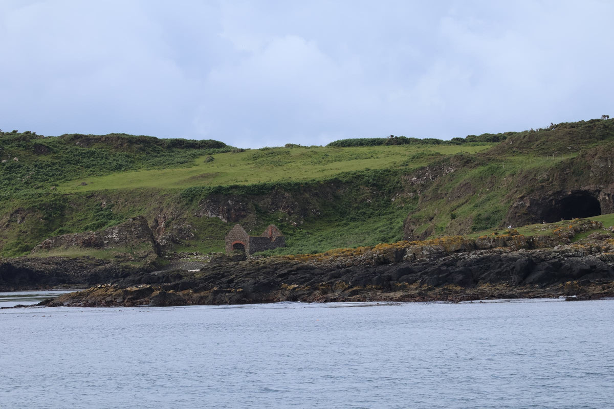 Coastal cliffs with a green clifftop above grey rock, a small stone ruin visible on the shore below.