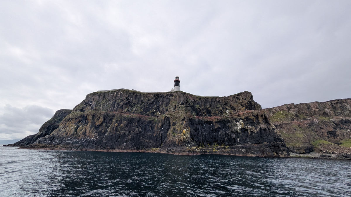 The East Light lighthouse standing on top of dark basalt cliffs on Rathlin, seen from the water under a flat grey sky.